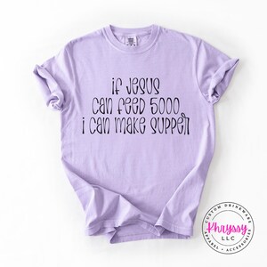 May include: A light purple t-shirt with the text "if Jesus can feed 5000 i can make supper" in black script. The shirt has rolled-up sleeves and a crew neck. A logo is visible in the bottom right corner.