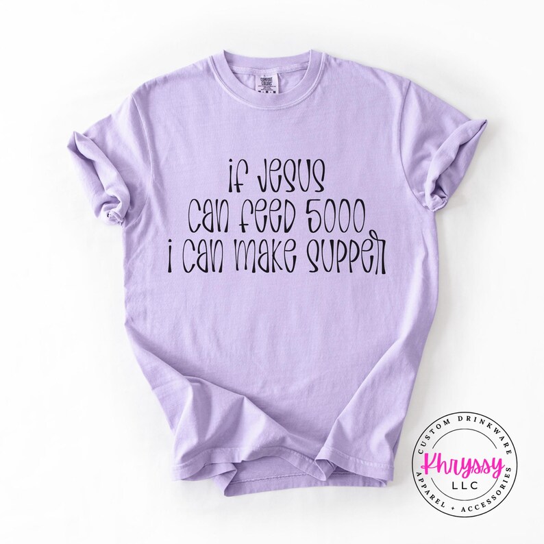 May include: A light purple t-shirt with the text "if Jesus can feed 5000 i can make supper" in black script. The shirt has rolled-up sleeves and a crew neck. A logo is visible in the bottom right corner.