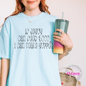 May include: Light blue t-shirt with the text "If Jesus can feed 5000, I can make supper" in black script. A teal tumbler with a straw is held in the hand. The tumbler has a gradient of pink, purple, and yellow.