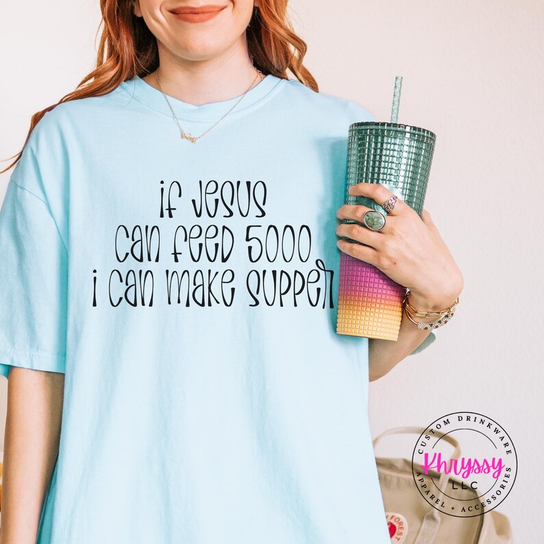 May include: Light blue t-shirt with the text "If Jesus can feed 5000, I can make supper" in black script. A teal tumbler with a straw is held in the hand. The tumbler has a gradient of pink, purple, and yellow.