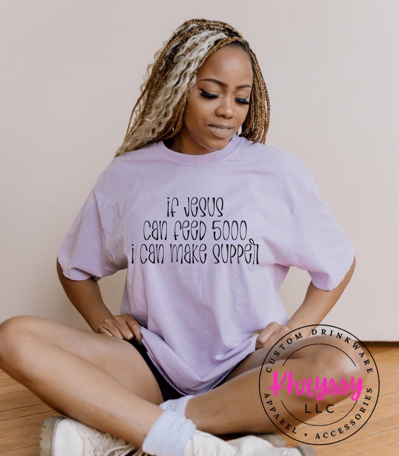 May include: A light purple t-shirt with the text "if Jesus can feed 5000 i can make supper" in black script. The shirt is worn by a person sitting on a wooden floor. A circular logo is visible.