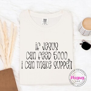 May include: Cream-colored t-shirt with the text "if Jesus can feed 5000, I can make supper" in black script. The shirt has a crew neck and a tag that says "Comfort Colors". A coffee cup, sunglasses, and a notebook are also visible.
