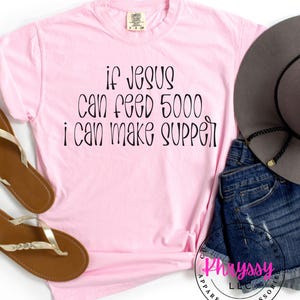May include: A light pink t-shirt with the text "if Jesus can feed 5000 I can make supper" in black script. The shirt is paired with denim shorts, a gray hat, and brown sandals.