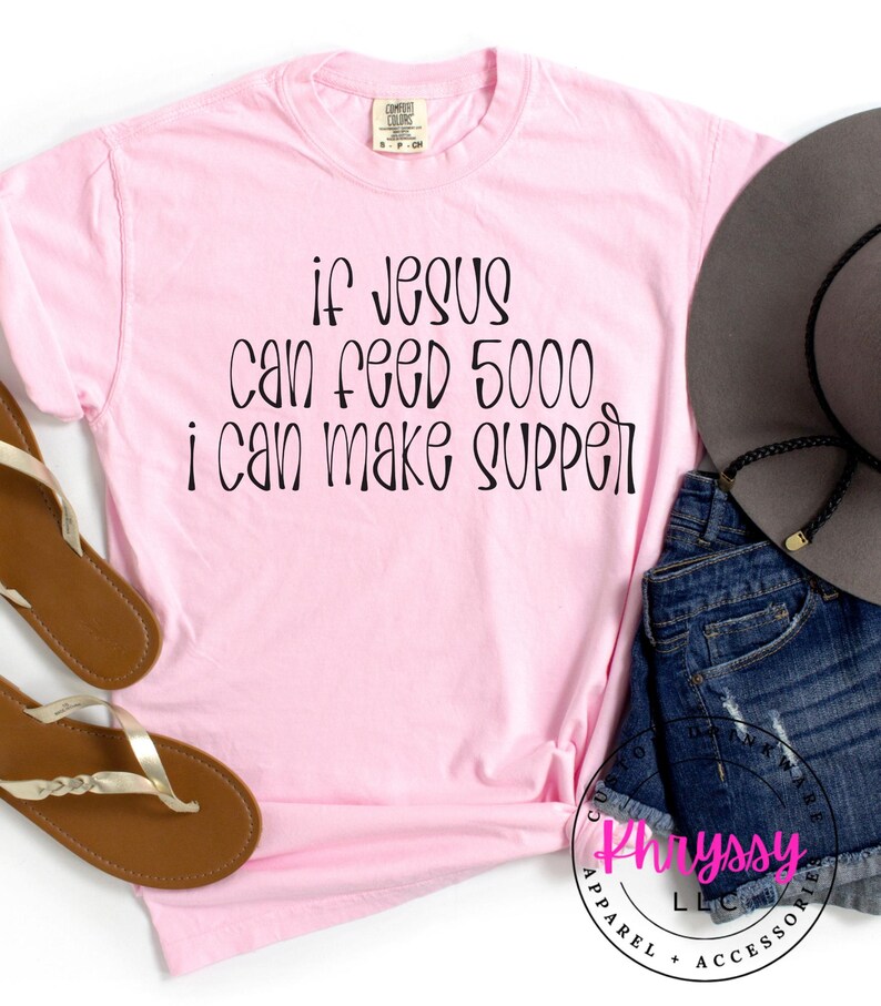 May include: A light pink t-shirt with the text "if Jesus can feed 5000 I can make supper" in black script. The shirt is paired with denim shorts, a gray hat, and brown sandals.