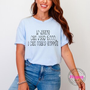 May include: A light blue t-shirt with the text "If Jesus Can Feed 5000, I Can Make Supper" in black script. The shirt is worn with blue jeans.