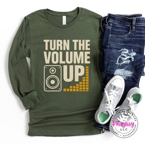 以下が含まれることがあります： 「TURN THE VOLUME UP」の文字とスピーカーのグラフィックが入ったオリーブグリーンの長袖シャツ。黄色いサウンドウェーブのグラフィックも特徴です。このシャツは、ブルージーンズとグレーのスニーカーと組み合わせています。