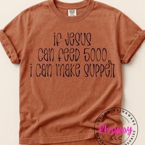 May include: A rust-colored t-shirt with the text "if Jesus can feed 5000, I can make supper" in black script. The shirt has rolled-up sleeves and a crew neck. The brand name is visible on the shirt.