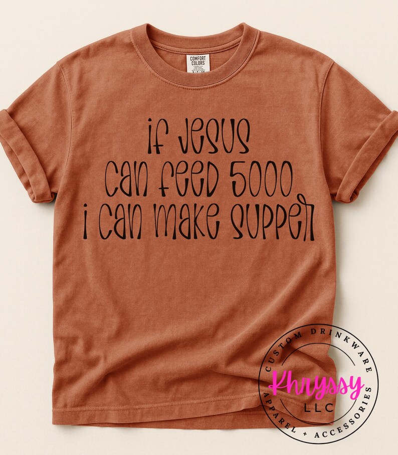 May include: A rust-colored t-shirt with the text "if Jesus can feed 5000, I can make supper" in black script. The shirt has rolled-up sleeves and a crew neck. The brand name is visible on the shirt.