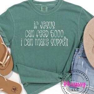 May include: A teal long-sleeve shirt with the text "if Jesus can feed 5000, I can make supper" in white script. The shirt is knotted at the waist. Accessories include sandals and a hat.