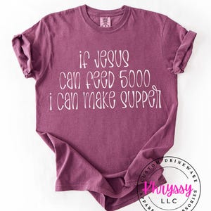 May include: A maroon t-shirt with the text "if Jesus can feed 5000 i can make supper" in white. The shirt has a crew neck and short sleeves. The bottom right corner has a circular logo with text.