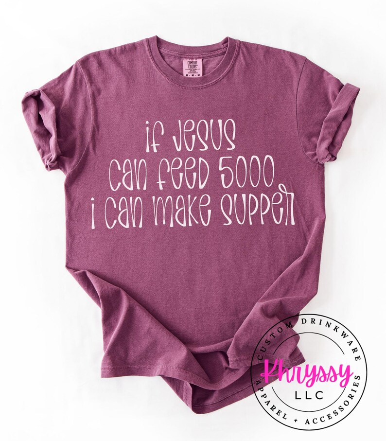 May include: A maroon t-shirt with the text "if Jesus can feed 5000 i can make supper" in white. The shirt has a crew neck and short sleeves. The bottom right corner has a circular logo with text.
