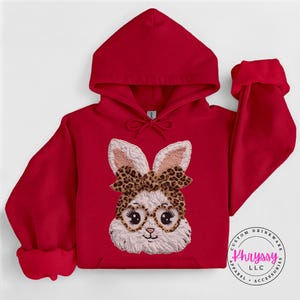 Può includere: Una felpa con cappuccio rossa con un viso da coniglio ricamato con occhiali e un fiocco leopardato. Il coniglio ha la pelliccia bianca e le orecchie rosa. La felpa ha un cappuccio con coulisse e polsini. Il logo "Khryssy LLC" è in basso a destra.