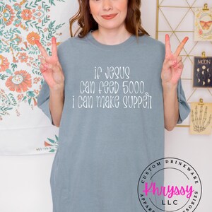 May include: A light blue t-shirt with the text "if Jesus can feed 5000, I can make supper" in white. The shirt also has a circular logo that says "Khryssy LLC Apparel + Accessories".
