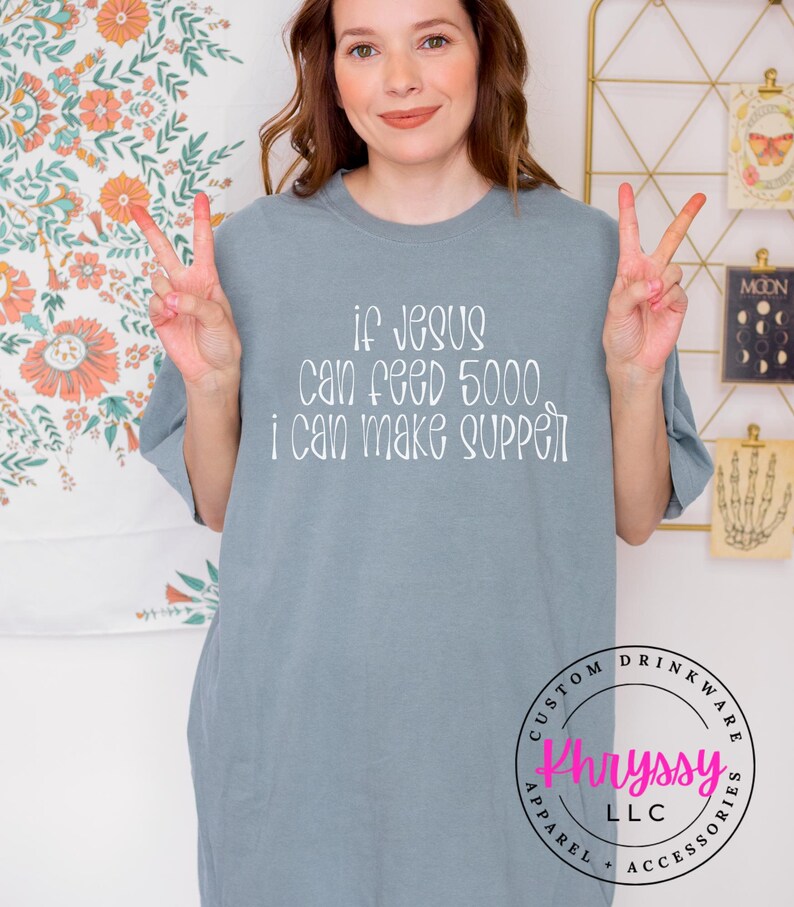 May include: A light blue t-shirt with the text "if Jesus can feed 5000, I can make supper" in white. The shirt also has a circular logo that says "Khryssy LLC Apparel + Accessories".