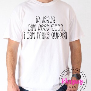 May include: White t-shirt with the text "If Jesus can feed 5000, I can make supper" in black script. The shirt is a casual style, suitable for everyday wear. The shirt also has a logo on the bottom right.