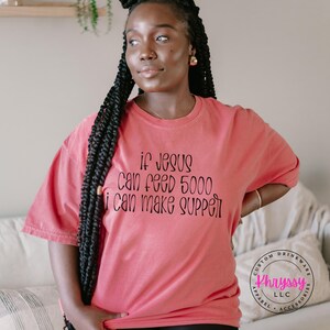 May include: A coral-colored t-shirt with the text "if Jesus can feed 5000 i can make supper" in black script. The shirt is a relaxed fit and has short sleeves. A custom drinkware logo is visible in the bottom right corner.
