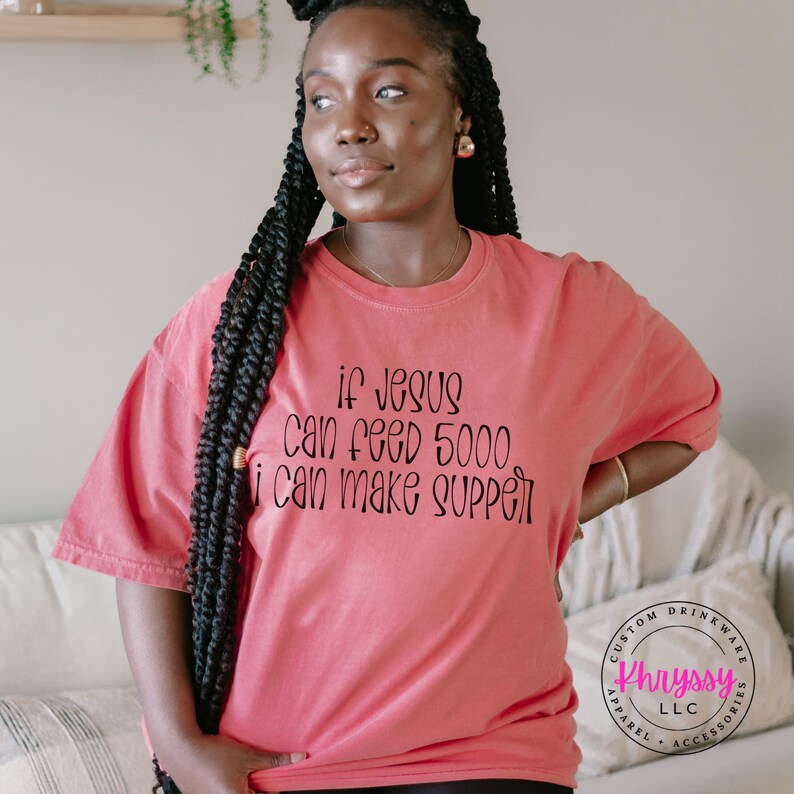May include: A coral-colored t-shirt with the text "if Jesus can feed 5000 i can make supper" in black script. The shirt is a relaxed fit and has short sleeves. A custom drinkware logo is visible in the bottom right corner.