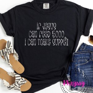 May include: Black t-shirt with white text that reads "If Jesus can feed 5000, I can make supper." The shirt is paired with denim shorts and sandals with black and white striped straps. The shirt is from Khryssy Apparel + Accessories.