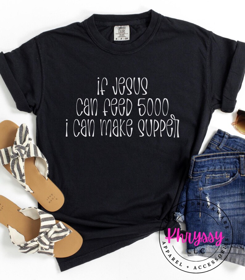 May include: Black t-shirt with white text that reads "If Jesus can feed 5000, I can make supper." The shirt is paired with denim shorts and sandals with black and white striped straps. The shirt is from Khryssy Apparel + Accessories.