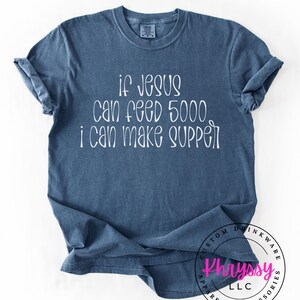 May include: A blue t-shirt with the text "if Jesus can feed 5000, I can make supper" in white script. The shirt has a crew neck and short sleeves. The bottom right corner has a circular logo.