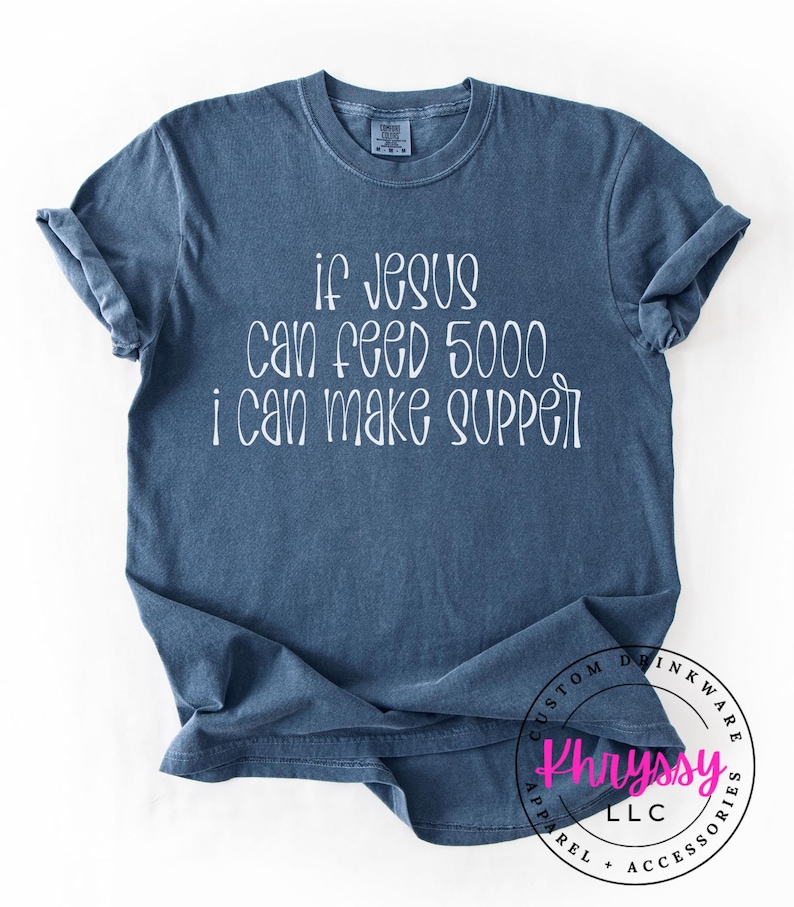 May include: A blue t-shirt with the text "if Jesus can feed 5000, I can make supper" in white script. The shirt has a crew neck and short sleeves. The bottom right corner has a circular logo.
