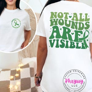 May include: White t-shirt with green text and a green ribbon design. The front of the shirt has a design that says "Mental Health Awareness". The back of the shirt says "Not All Wounds Are Visible".