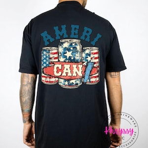 May include: Black t-shirt featuring a patriotic design with the word "AMERI" arched across the top. Below, three stylized cans are depicted with red, white, and blue star and stripe patterns, and the word "CAN" is centered.