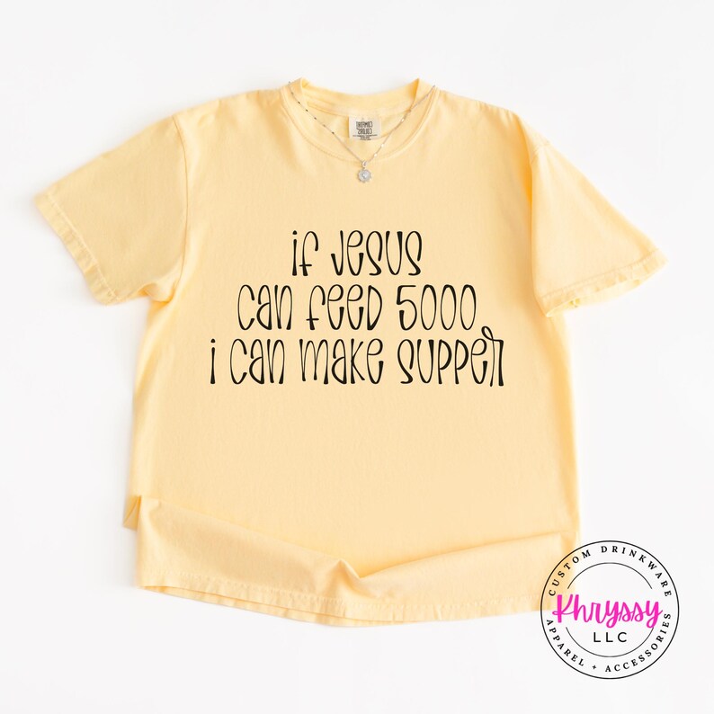 May include: A light yellow t-shirt with the text "if Jesus can feed 5000, I can make supper" in black. The shirt has short sleeves and a crew neck. A small necklace is visible. The bottom right corner has a circular logo.