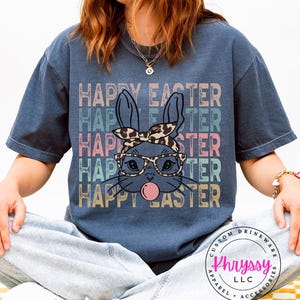 May include: A navy blue t-shirt with the words "HAPPY EASTER" stacked vertically in pastel colors. The shirt features a cartoon bunny wearing glasses and a leopard print headband, blowing a bubble.