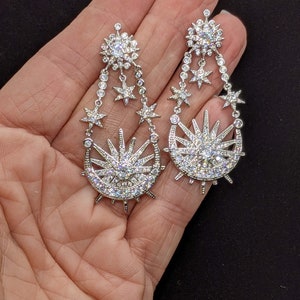 May include: A pair of silver dangle earrings with a crescent moon and sun design. The earrings are adorned with clear cubic zirconia stones.
