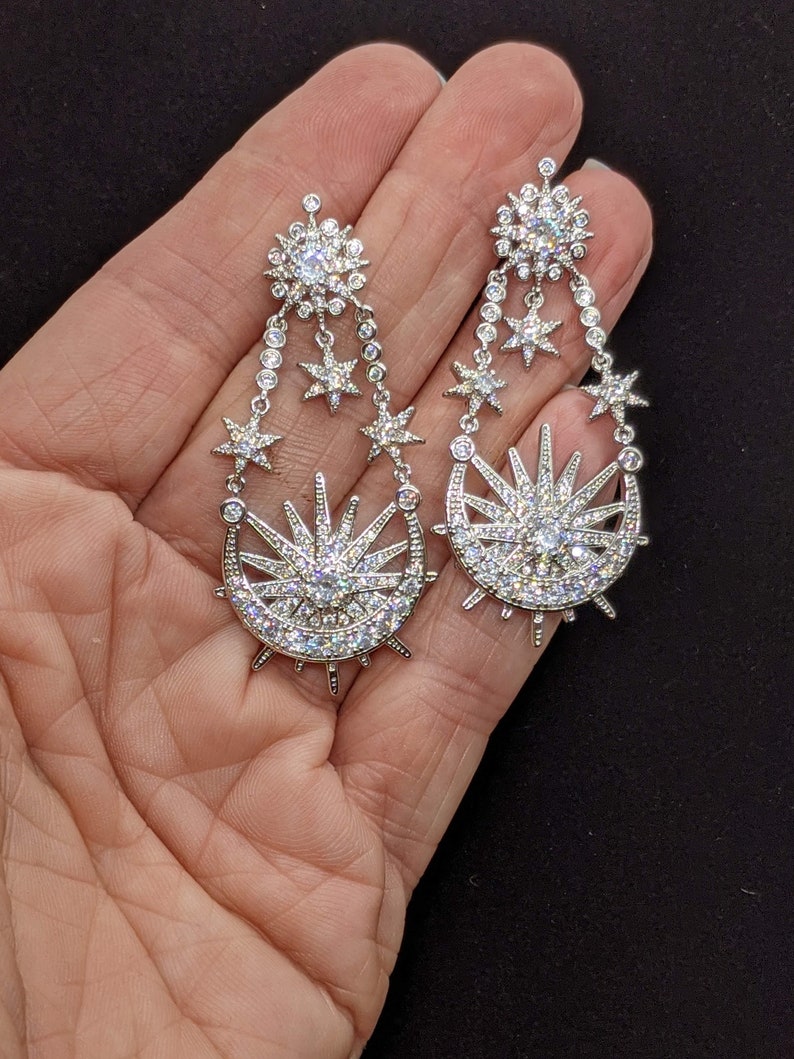 May include: A pair of silver dangle earrings with a crescent moon and sun design. The earrings are adorned with clear cubic zirconia stones.