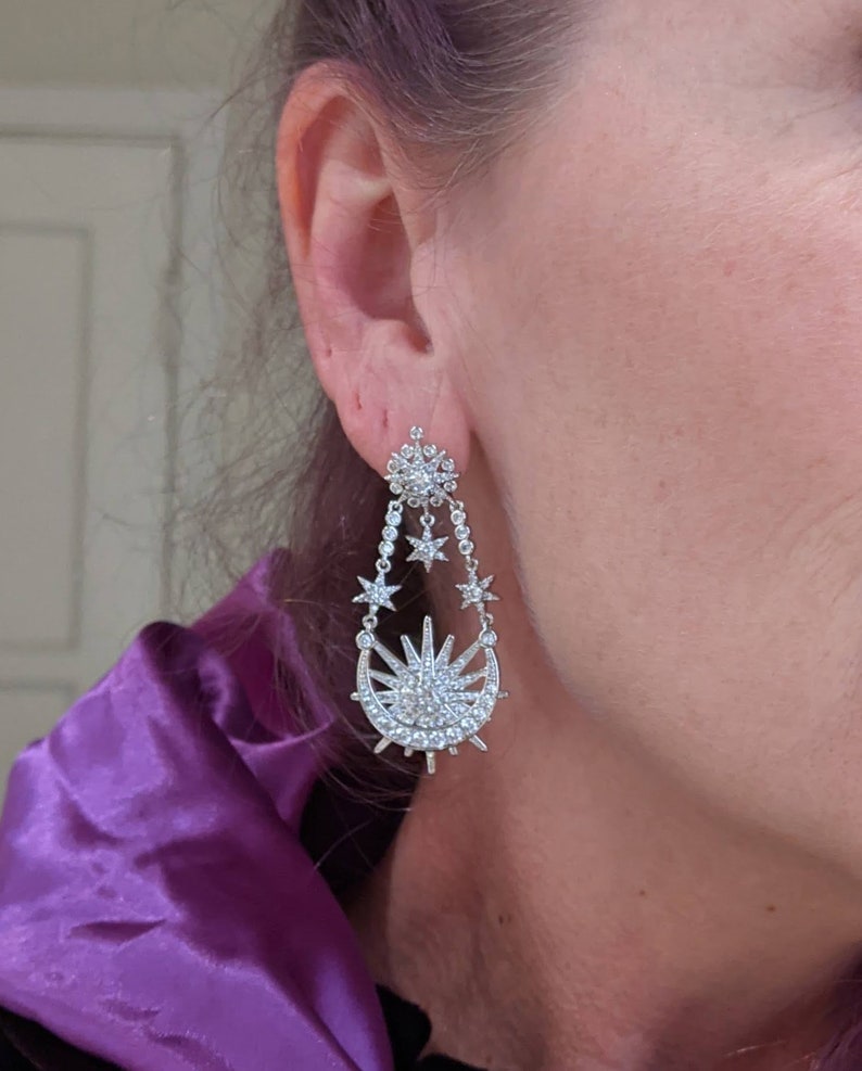 May include: Silver dangle earrings with a crescent moon and sun design, adorned with sparkling clear crystals.