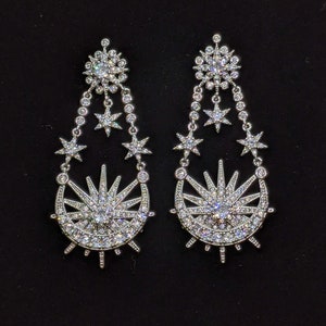 May include: A pair of silver earrings with a crescent moon and sun design. The earrings are adorned with sparkling clear crystals.