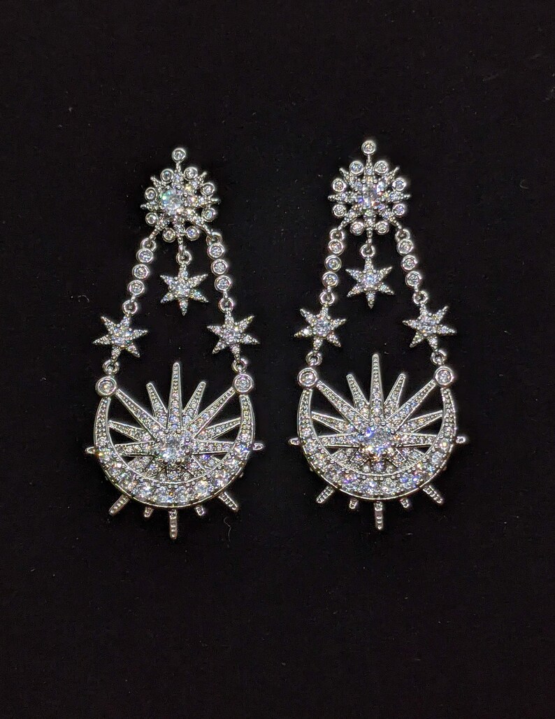 May include: A pair of silver earrings with a crescent moon and sun design. The earrings are adorned with sparkling clear crystals.