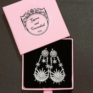 May include: A pair of silver dangle earrings with a crescent moon and star design. The earrings are in a pink box with the text "Queen and Cavendish 1774".