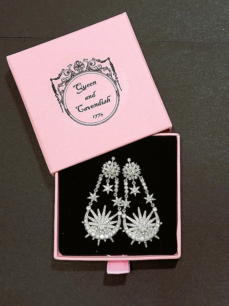 May include: A pair of silver dangle earrings with a crescent moon and star design. The earrings are in a pink box with the text "Queen and Cavendish 1774".