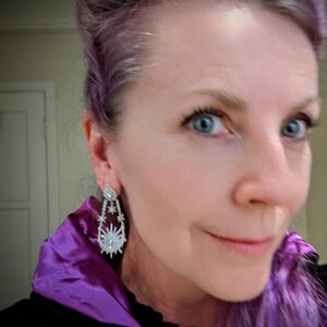 May include: A woman with purple hair wearing a black velvet jacket with a purple satin collar. She is wearing large silver dangle earrings with a starburst design.