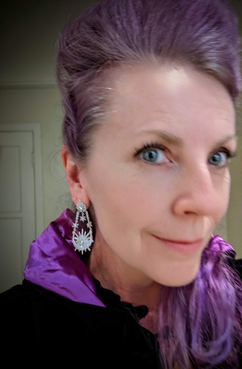 May include: A woman with purple hair wearing a black velvet jacket with a purple satin collar. She is wearing large silver dangle earrings with a starburst design.