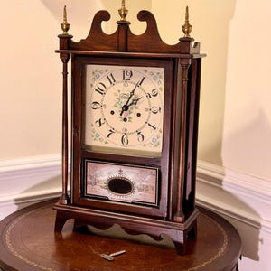 May include: A dark wood antique clock with a cream-colored face featuring black Roman numerals and delicate floral accents. The clock has a rectangular shape with a decorative top and a small landscape painting on the lower section. A key rests on the table.