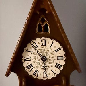 May include: A brown wooden clock in a church-like shape. The clock face is white with black Roman numerals and the words "Hark, Prepare". The clock has a pendulum and a decorative cross on top.