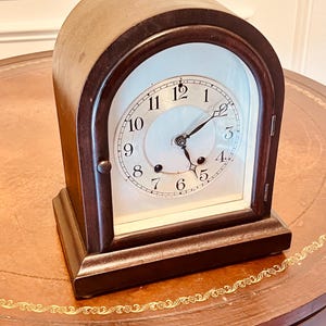 May include: A dark brown wooden mantel clock with a white face and black hands. The clock has Roman numerals and is set on a brown table with gold trim.