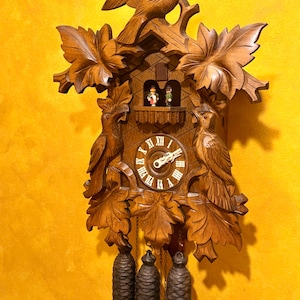 May include: A wooden cuckoo clock with intricate carvings of birds, leaves, and a small house. The clock face has Roman numerals, and three pinecone-shaped weights hang below. The clock is made of brown wood and has a detailed design.