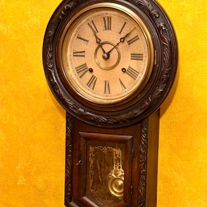 May include: Antique wooden wall clock with a round face and Roman numerals. The clock has a dark brown, ornate frame and a pendulum visible through a glass door. The clock is mounted on a yellow wall.