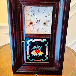 May include: Antique wooden clock with a painted face and a key. The clock has Roman numerals and a floral design. The clock is in a dark wood frame.
