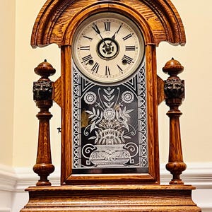 May include: Antique wooden clock with a Roman numeral face and a decorative floral design on the glass. The clock has a dark wood finish and ornate details.