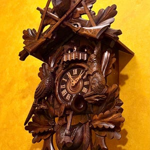 May include: A wooden cuckoo clock with a deer head and antlers at the top. The clock face has Roman numerals and is flanked by a bird and a squirrel. Decorative leaves and pinecone weights complete the design.