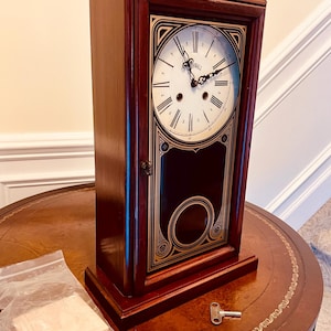May include: A wooden wall clock with a black and gold face and a key for winding. The clock has Roman numerals and a small, white dial with the word "Westwall" on it.