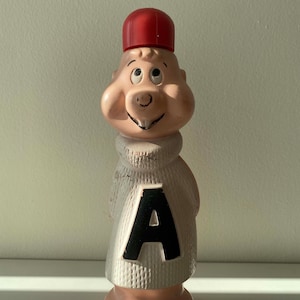 Puede incluir: Una figurita de juguete de plástico vintage de un personaje de dibujos animados que lleva un sombrero rojo y un suéter blanco con una letra 'A' negra en la parte delantera. El personaje tiene una cara rosa y un cuerpo blanco.