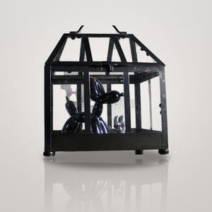 Könnte beinhalten: Ein schwarzes, hausförmiges Terrarium aus Metall mit Glasscheiben, das eine glänzend schwarze Ballonhund-Figur und eine kleinere silberne Ballonhund-Figur enthält. Die Struktur hat ein Spitzdach und einen rechteckigen Sockel mit vier Füßen.