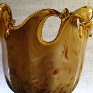 May include: A hand-blown amber glass vase featuring a swirling marbled pattern with deep brown and golden hues. The vessel has an irregular, wavy rim with a prominent cutout opening near the top, creating a fluid and organic silhouette.